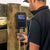 Cattle Rancher in a black shirt stands beside a wooden fence post holding a smartphone, while a digital livestock scale with an orange border is mounted on the post. The scale displays a reading of 478, and a rope hangs from it.